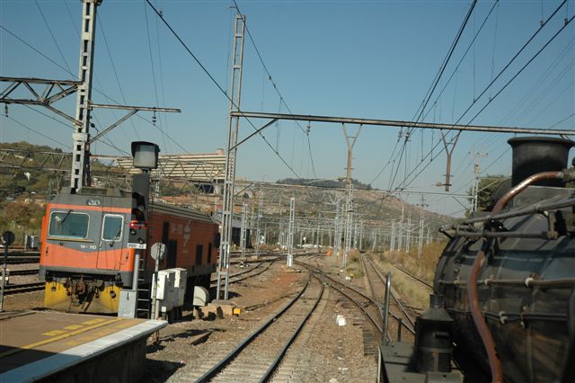 As the steam-hauled "Tshwane Xplorer" winds through the pointwork of Pretoria main station, it passes the class 14E electric unit used to haul the renowned Blue Train to Cape Town. This dedicated unit is stabled here at the platform end, awaitting the next Blue.