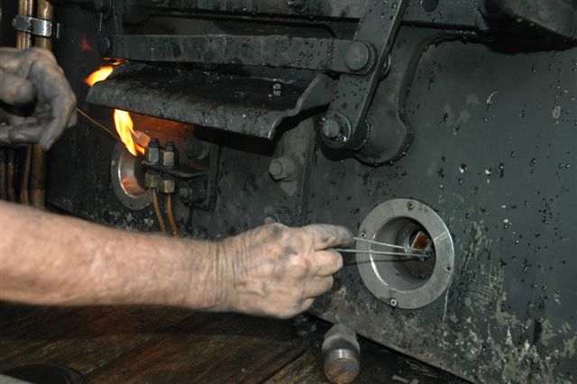 Here, he has placed the small mirror (right side), into the water space above the foundation ring, and provided light from the flare lamp to illuminate the area, so that he will be able to see if there is any obstruction or damage to the inner firebox, such as loose stays or scouring. This procedure will be repeated in all areas around the firebox area, both inside and outside the cab.